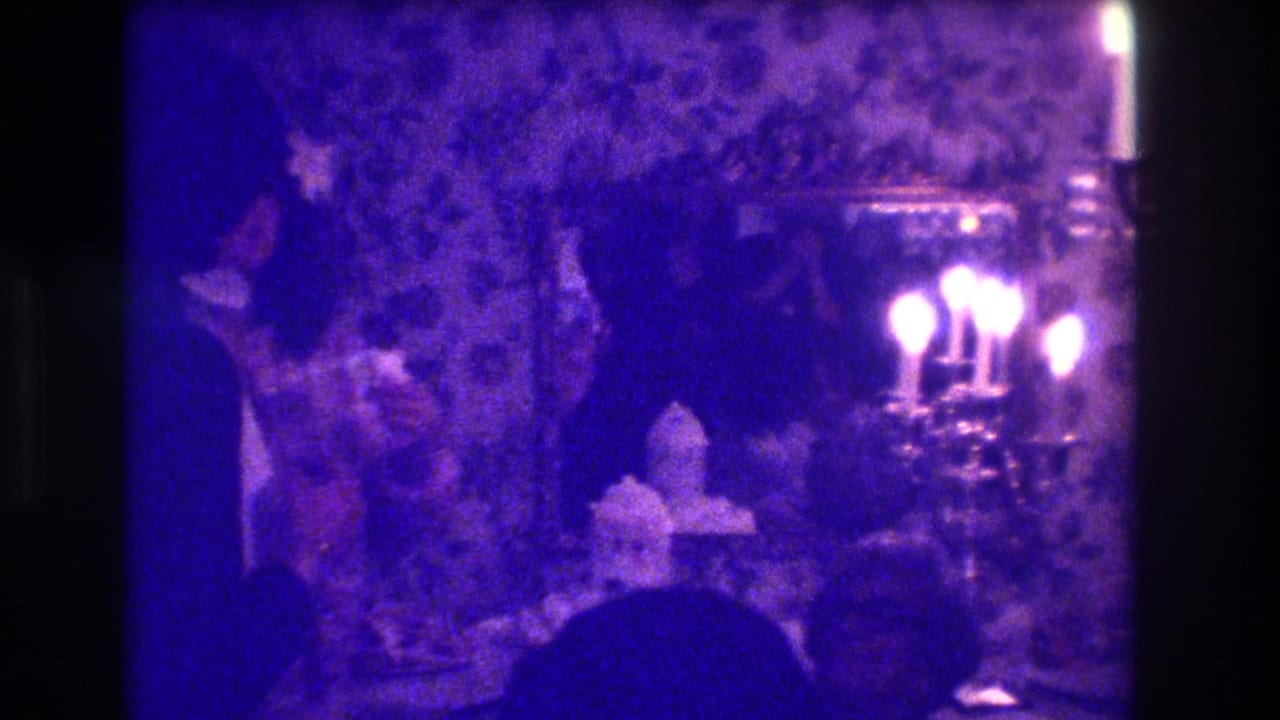 Two silhouetted figures stand near a table with cake and a bright candelabra reflected by a mirror.