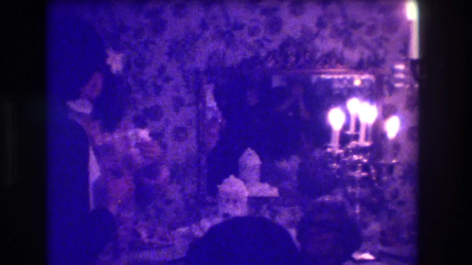 1973 Medford indoor celebration frame with two figures near a cake table, glowing candelabra, patterned wallpaper, and a mirror in low blue light.