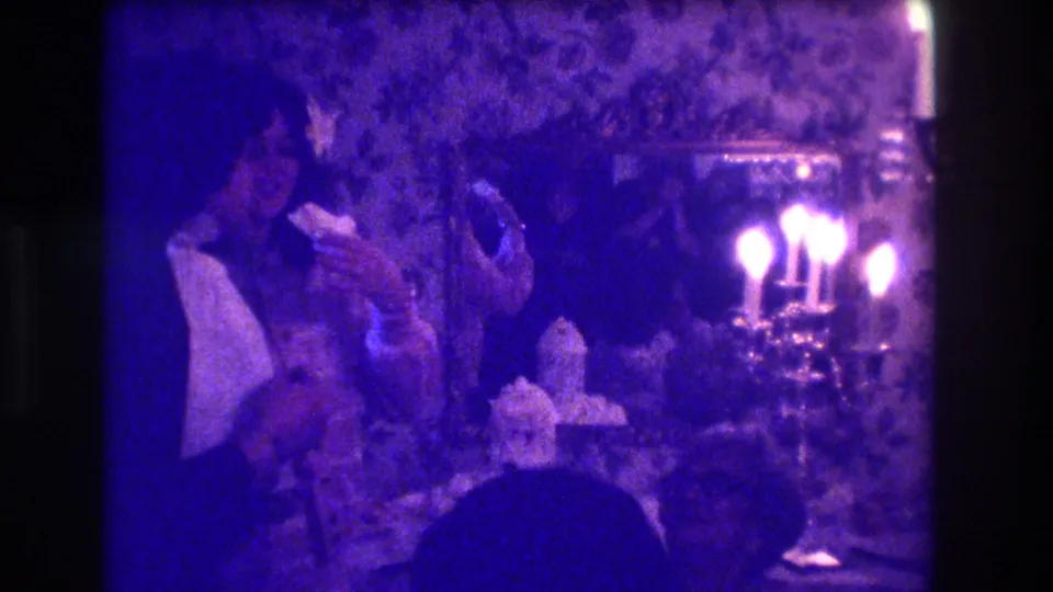 1973 Medford home-movie frame showing a person at a celebration table, cake shapes in front, candelabra lights, mirror, and wallpaper.