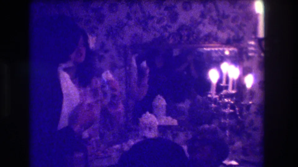 1973 Medford celebration frame with guests gathered around a cake table, bright candelabra flames, patterned wallpaper, and mirror in blue-purple film color.