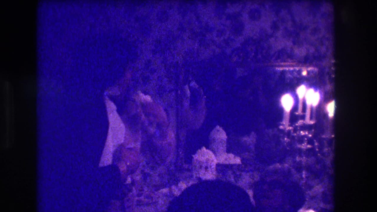 1973 Medford, New Jersey home-movie frame with formal guests, a candelabra, and patterned wallpaper under strong purple-blue film color.