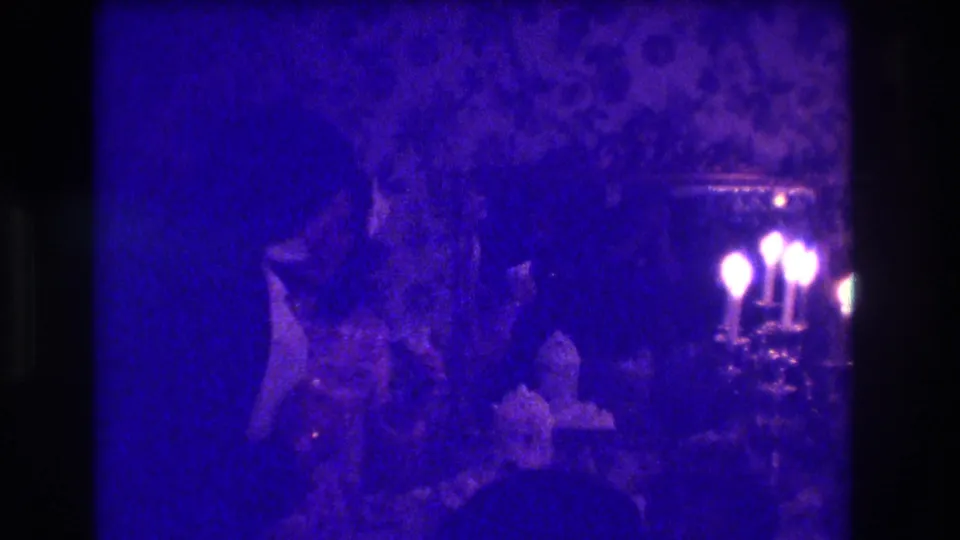 1973 Medford, New Jersey party frame showing adults in a dim private room, with candles glowing against floral wallpaper.