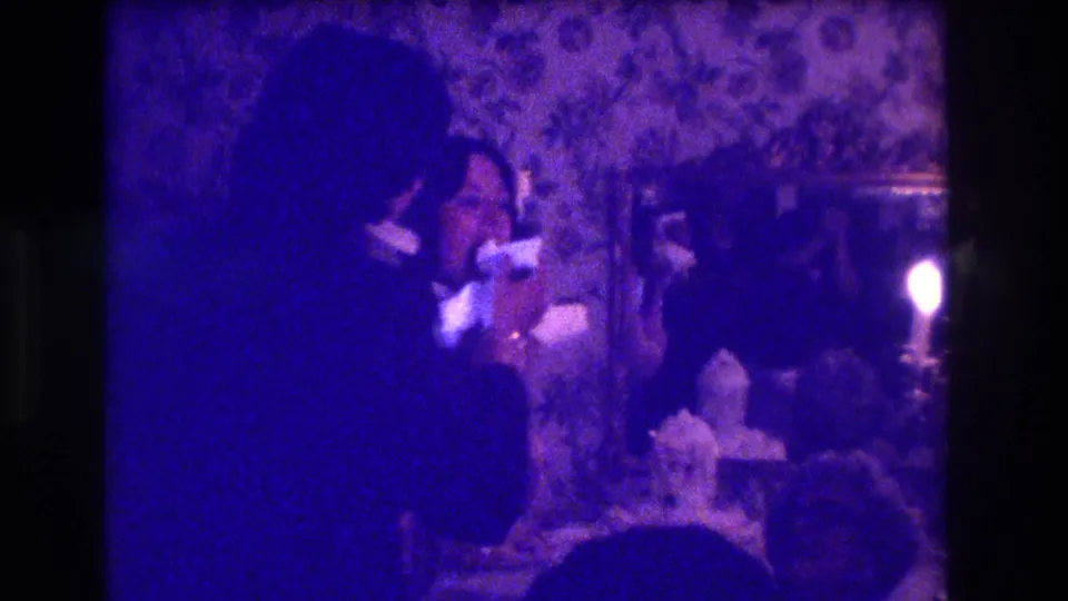 An adult appears to feed or share food with another person at a formal indoor event in this 1973 Medford home-movie frame.