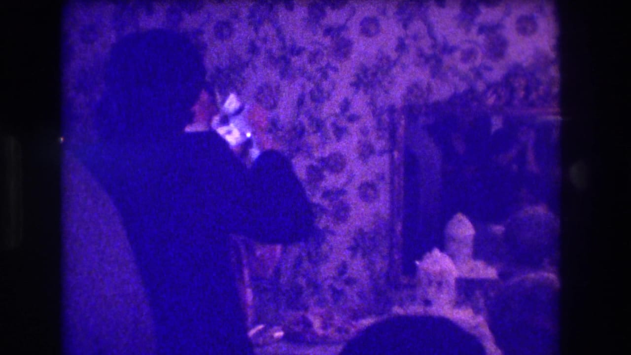 Purple-shifted 8mm frame of the groom turned toward the reception table, with cake, wallpaper, and wall decor visible.