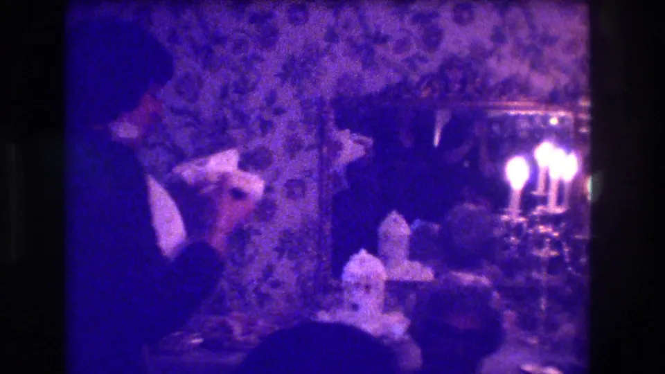Home-movie frame from the wedding reception showing the bride and groom near the cake, with candles and guests in a dim interior.