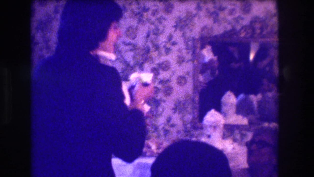 Suited man holds a glass in a dim room with floral wallpaper and seated guests.