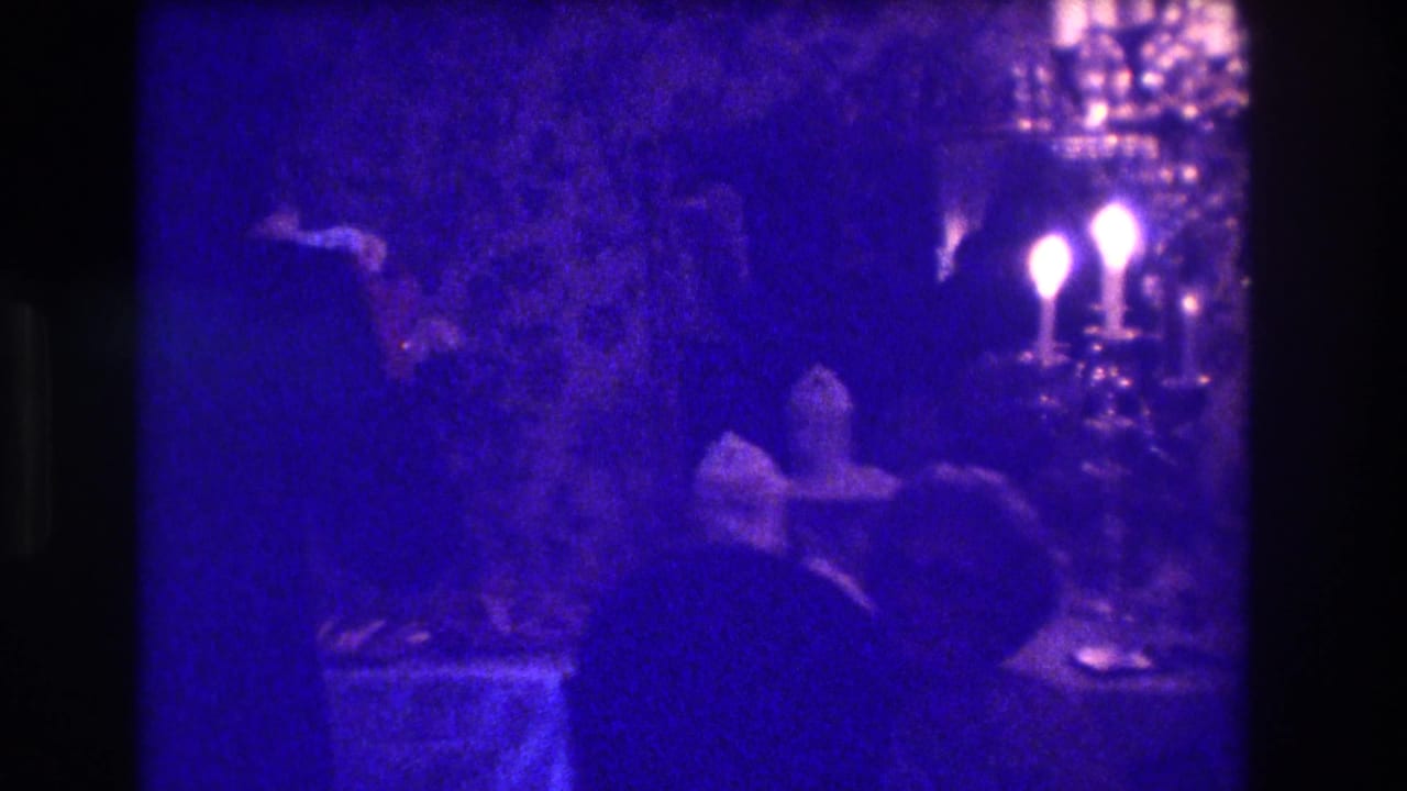 Blue-tinted 8mm frame from a Medford interior gathering shows shadowed guests near candle-style lights and table items.
