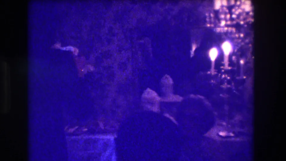 Blue-tinted 8mm frame from a Medford interior gathering shows shadowed guests near candle-style lights and table items.