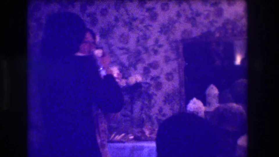 A suited man appears to drink from a glass during a 1973 Medford indoor gathering, with floral wallpaper and seated guests visible.