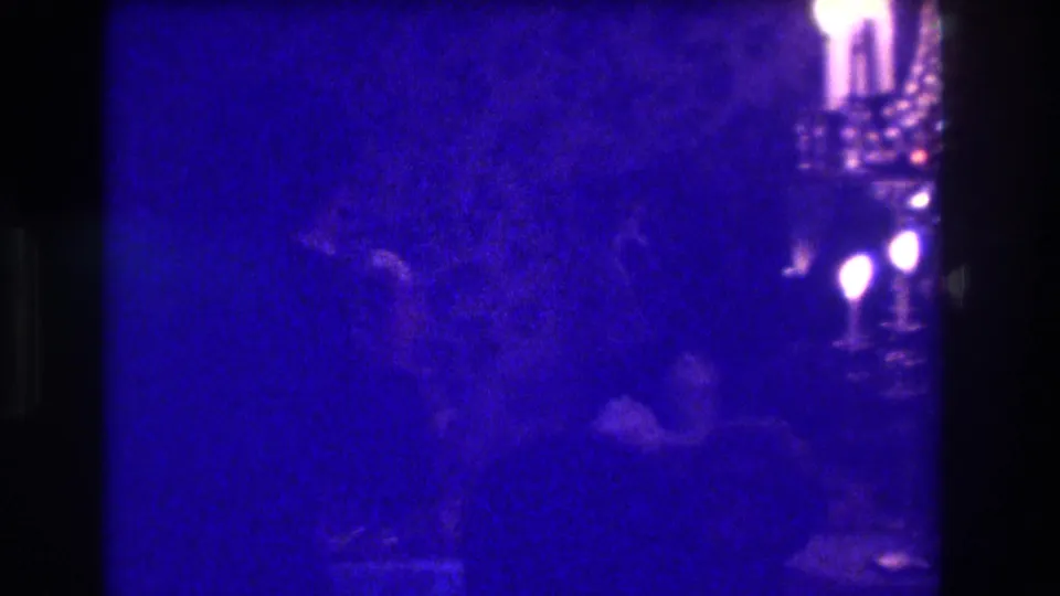 1973 Medford home-movie frame showing shadowed adult guests in a dim room with floral wallpaper and candlelit fixtures.