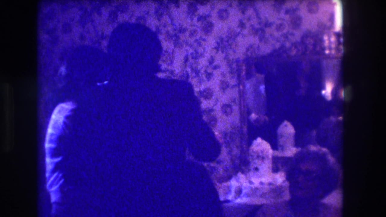 Two shadowed adults embrace beside floral wallpaper and a lit table area.