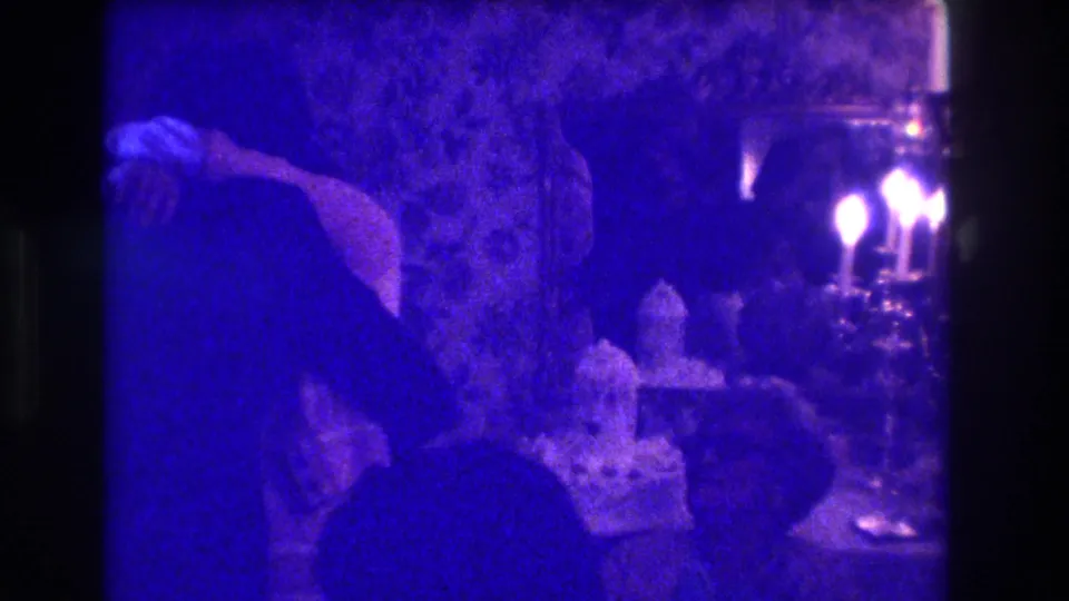 Blue-toned 1973 Medford home-movie frame showing party guests near candles, floral wallpaper, and tabletop decor.