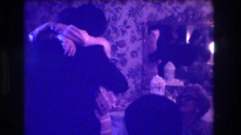1973 Medford indoor party frame showing two adults embracing while other guests sit nearby in a decorated private room.