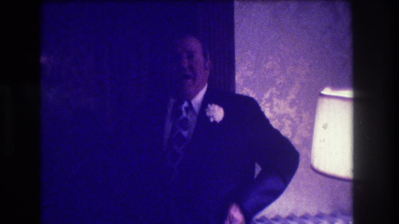 Suited man with a boutonniere stands beside a lamp in a dim interior.