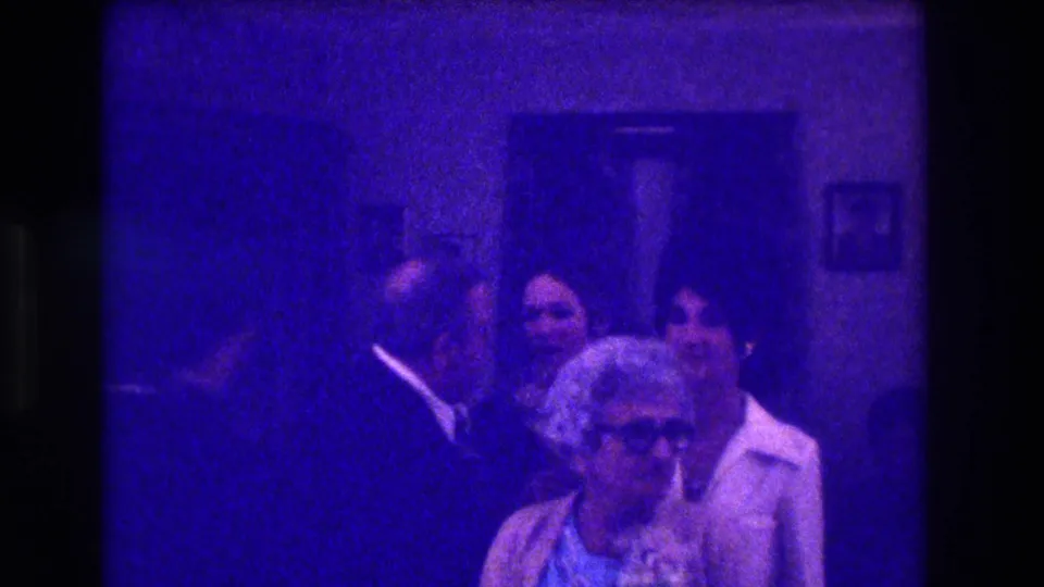 Medford, New Jersey, 1973 home-movie frame showing guests conversing behind an older woman in glasses in a blue-tinted room.