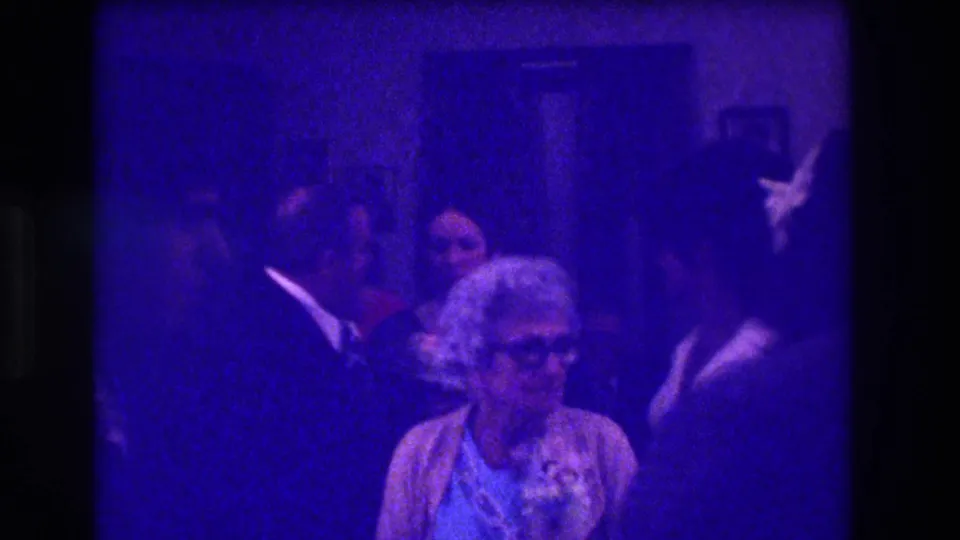 Medford, New Jersey, 1973 frame of an older woman with glasses in front of guests gathered in a private room.