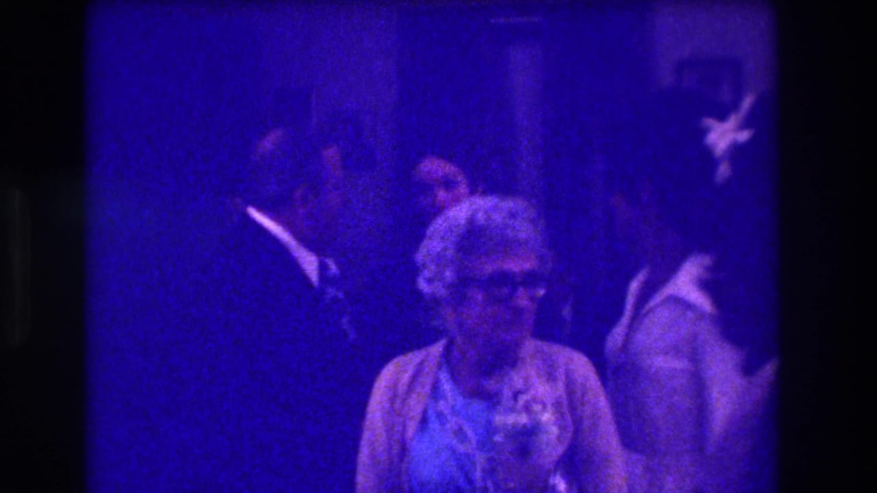 1973 Medford home-movie frame showing an older seated woman and nearby party guests under a strong purple-blue film cast.