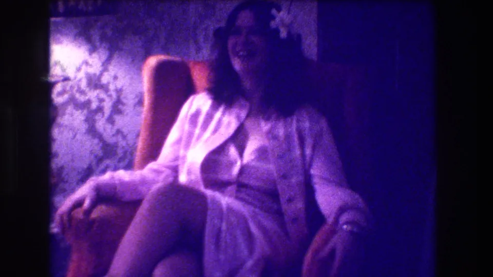 1973 Medford home-movie frame of a laughing woman in a light dress and cardigan seated in a patterned armchair.
