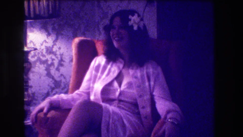 1973 Medford home-movie frame showing a laughing seated woman, flower in hair, lamp, armchair, and patterned interior wall.
