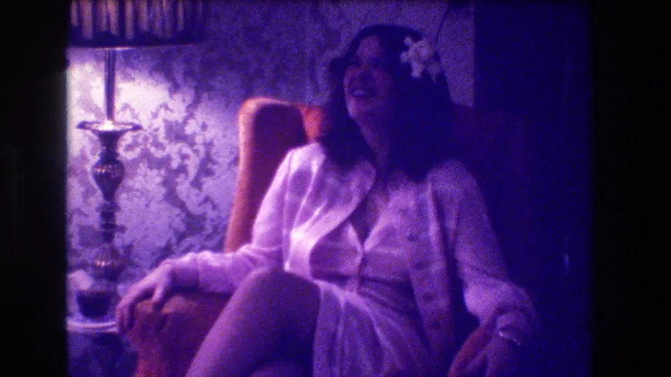 1973 Medford home-movie frame of a laughing woman seated in a private interior with lamp, wallpaper, and heavy color shift.