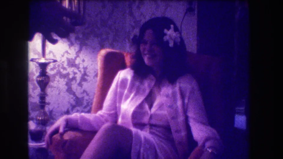 A woman seated in a domestic interior smiles while a hand enters the purple-lit 1973 Medford home-movie frame.