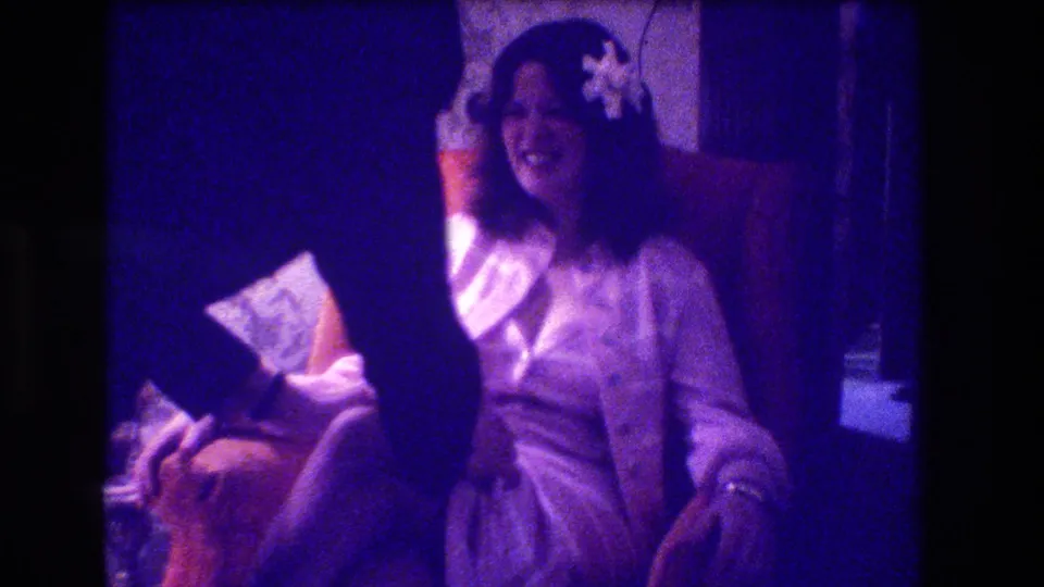 A smiling woman in a white robe or dress sits in a chair as another adult's leg or arm crosses the foreground.