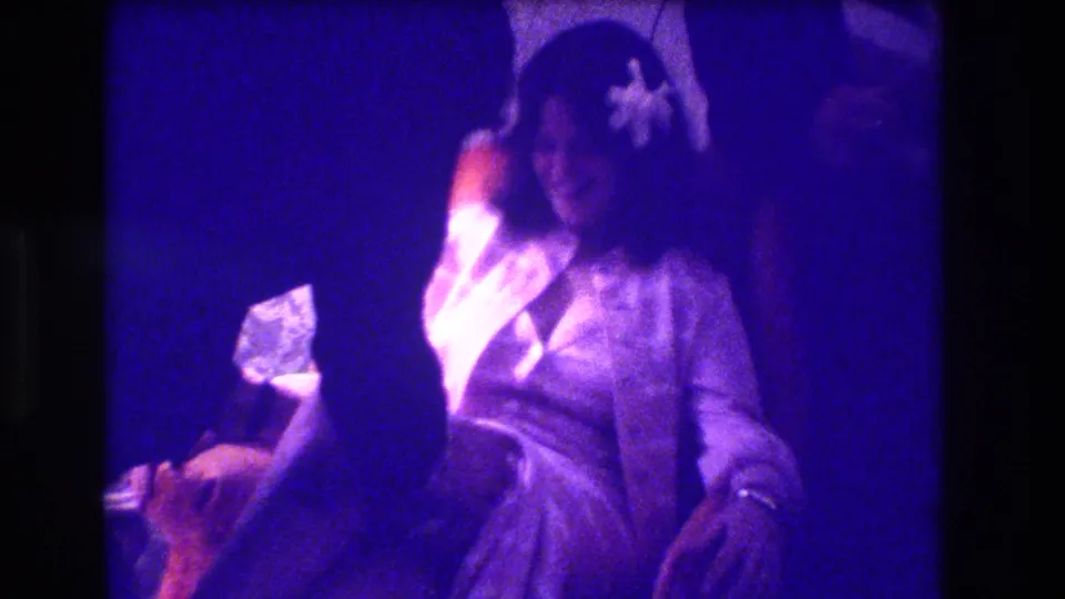 The Medford 1973 home-movie frame shows a seated woman partly obscured by another person in a dim purple-lit room.