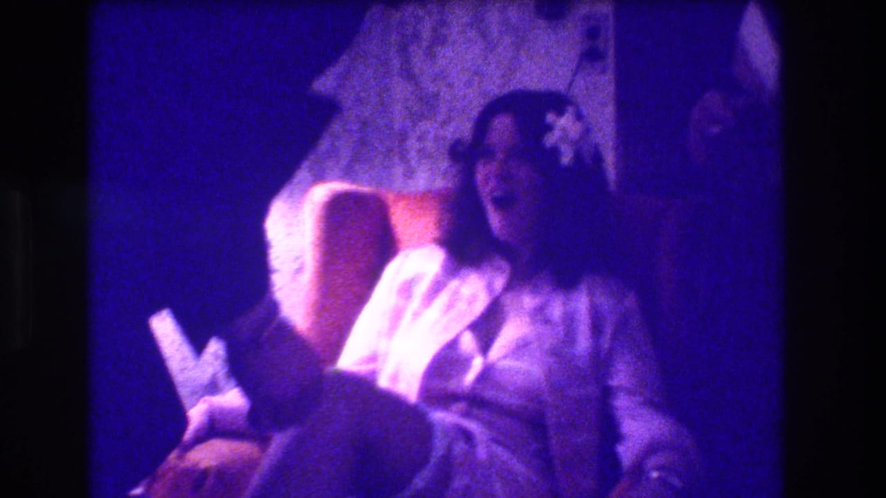 Seated bride in white laughing as a suited man leans near her leg indoors.
