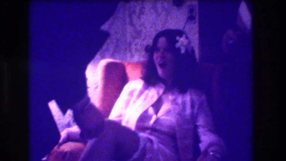 1973 Medford, New Jersey home-movie frame showing a seated bride laughing while a suited man leans in during a garter moment.
