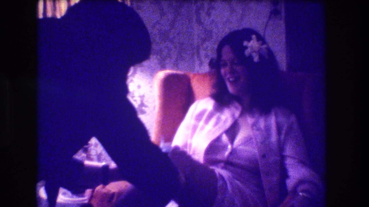 Bride in a white gown laughs from an upholstered chair while a silhouetted person leans in front of her.