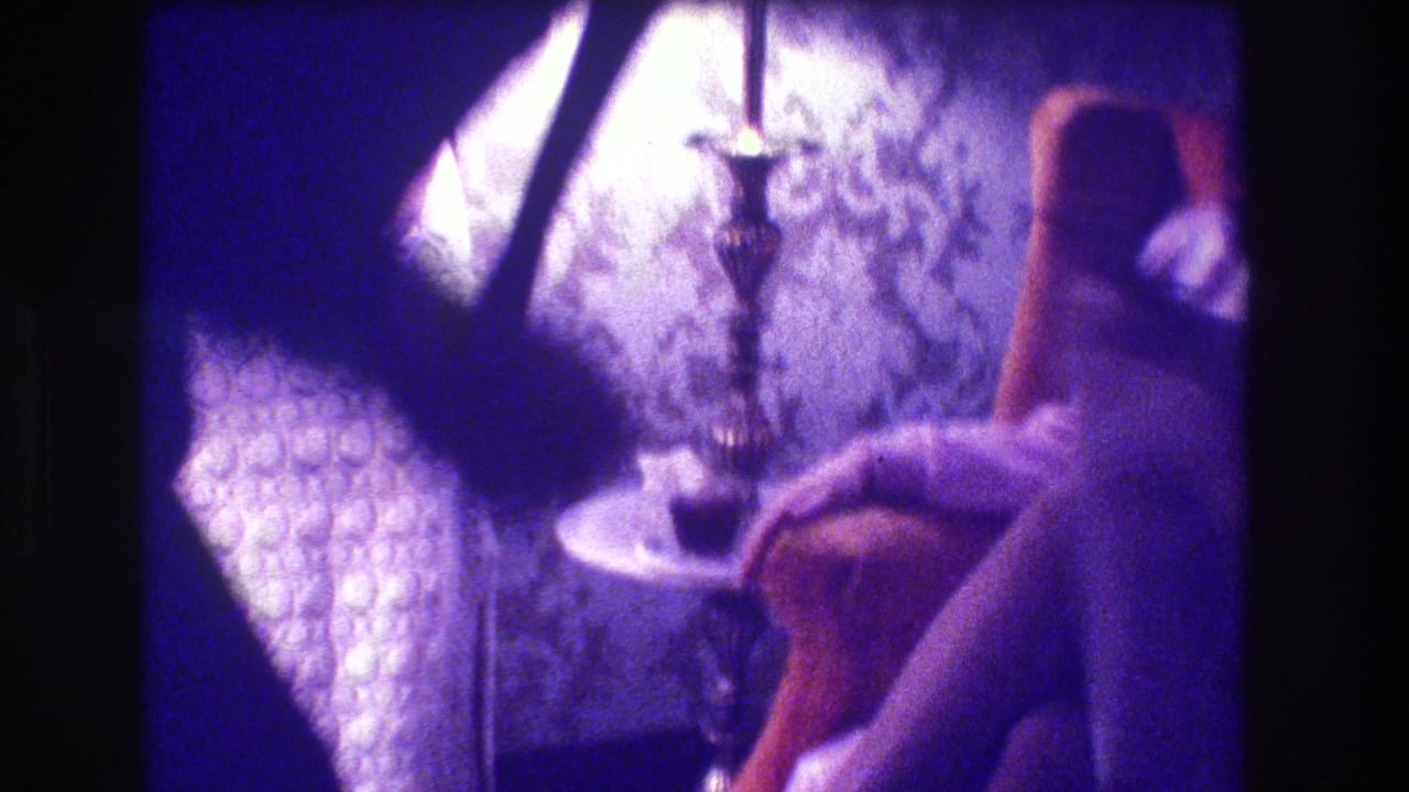 1973 Medford, New Jersey home-movie frame showing a seated attendee near patterned wallpaper, a small table, and a lamp or candleholder.