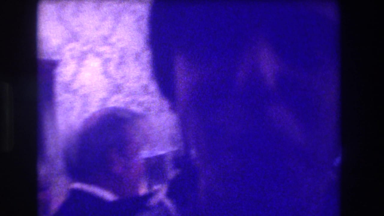 Blue-purple 8mm frame from the Medford indoor gathering, with a partly visible attendee and indistinct room furnishings.