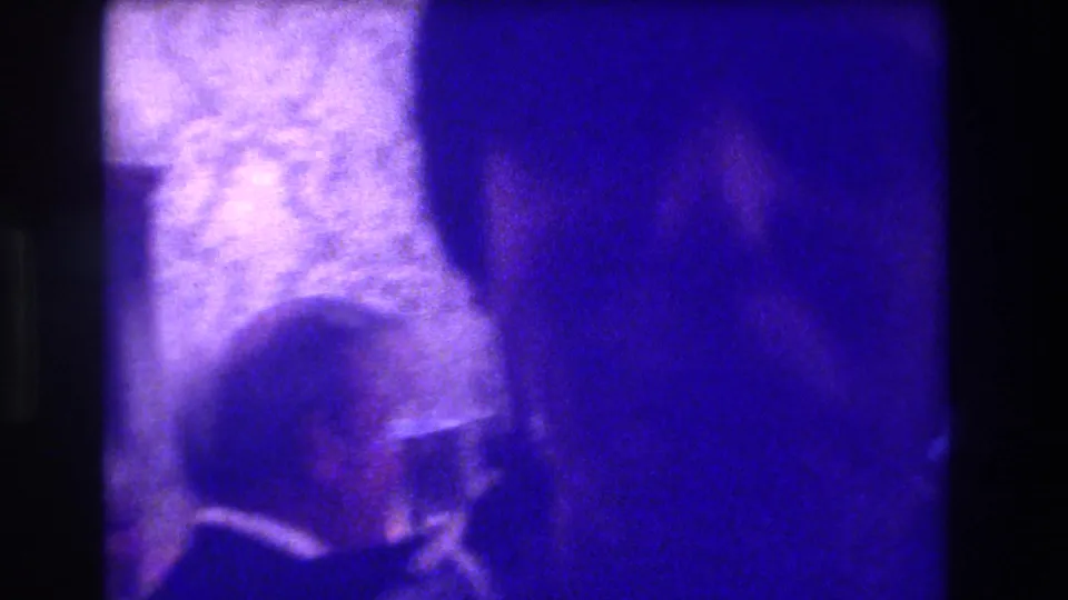 Blue-purple 8mm frame from the Medford indoor gathering, with a partly visible attendee and indistinct room furnishings.