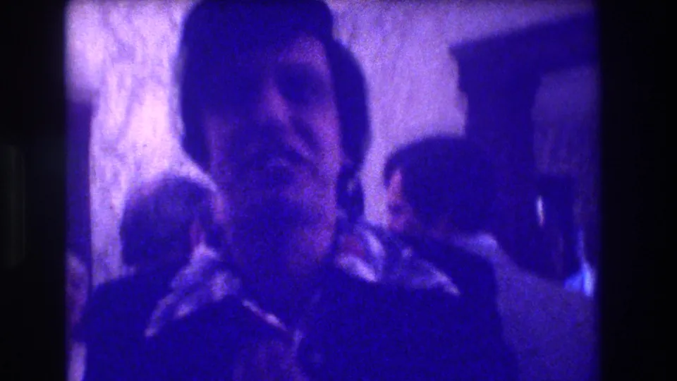 1973 Medford home-movie frame showing a man in dark clothing addressing the camera while attendees stand or sit behind him.