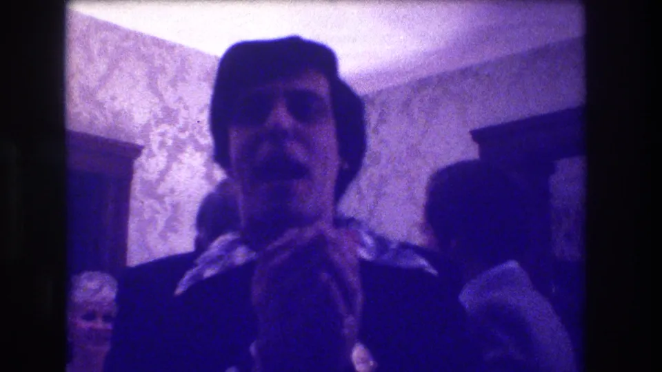 Medford, New Jersey 1973 frame showing a man in a dark suit and scarf or collar detail facing the camera among indoor attendees.
