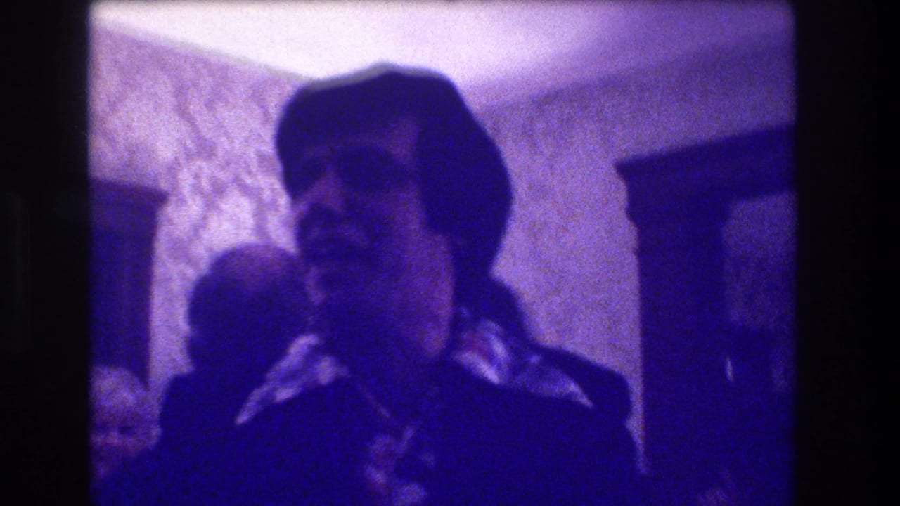 Young man in a dark jacket standing in a dim party room with patterned wallpaper.