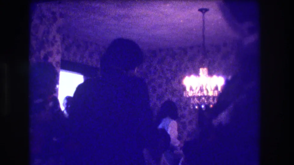 A crowded Medford party room shows silhouetted guests, patterned wallpaper and a lit chandelier in low-exposure 8mm footage.