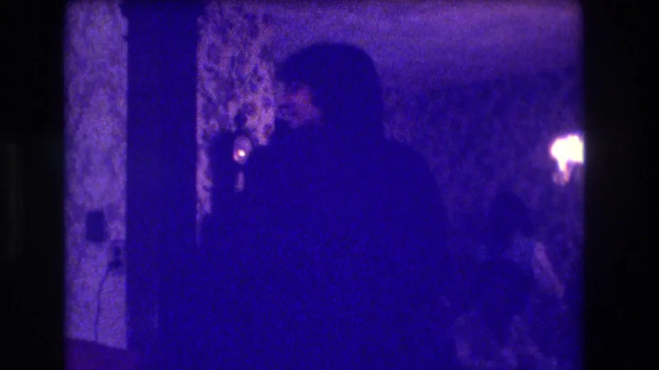1973 Medford, New Jersey home-movie frame showing adults silhouetted inside a dim dining room with patterned wallpaper and a small bright light.