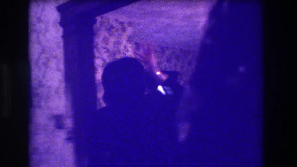 1973 Medford home-movie frame showing a dark figure in a dining-room interior, appearing to hold or point a small bright object.