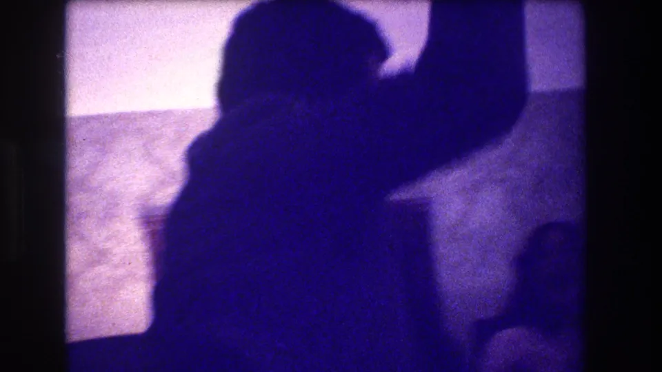 Low-light 1973 Medford home-movie frame with a close silhouetted adult raising an arm inside a private room.