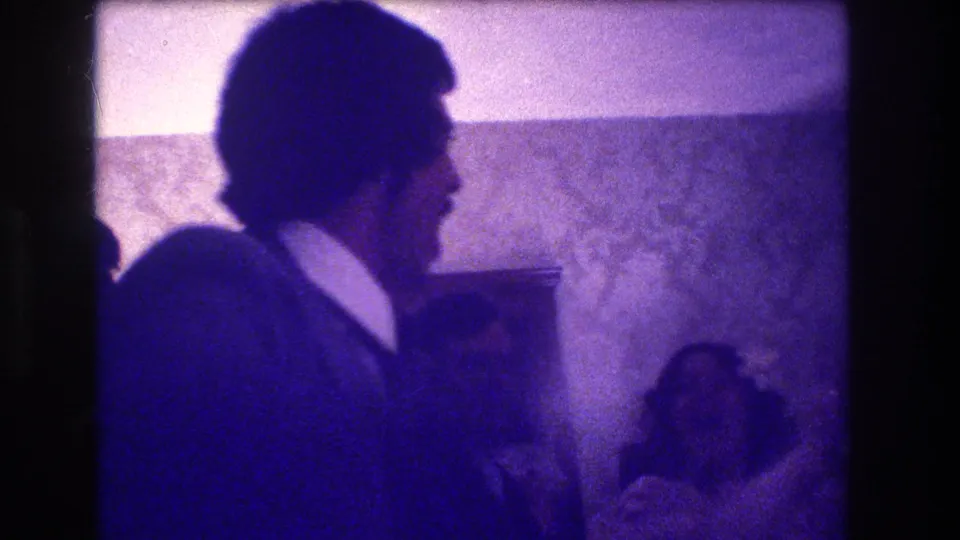 1973 Medford, New Jersey home-movie frame showing a man in profile clapping while other guests sit nearby in a private interior.