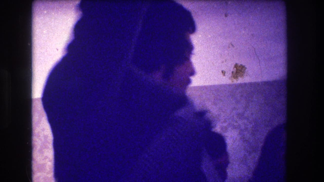 1973 Medford, New Jersey home-movie frame with adults leaning together in profile during an indoor party or family gathering.