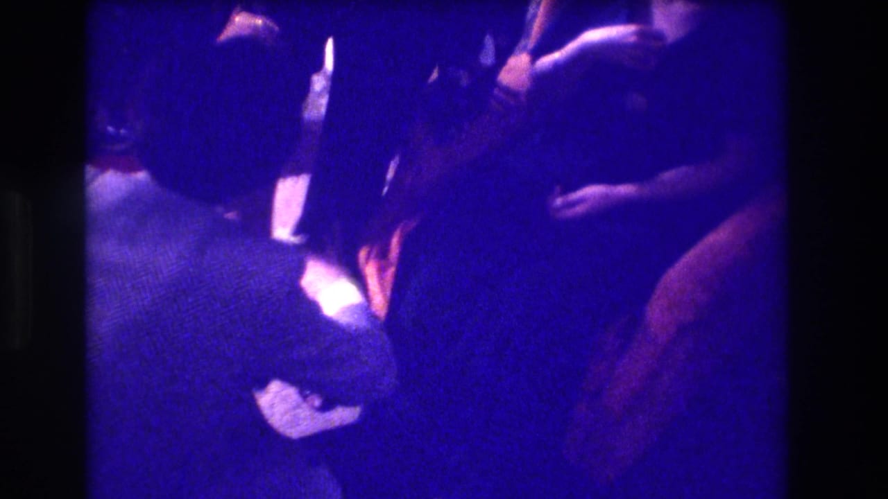 Blue-lit home-movie frame of adults close together in a dim private interior.