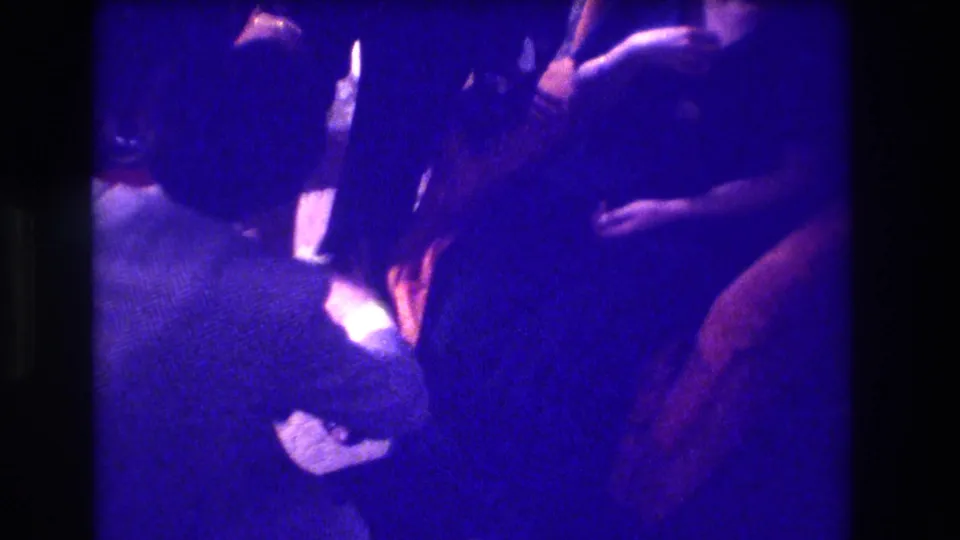 1973 Medford, New Jersey 8mm frame showing adults leaning close together under heavy blue-purple interior light.