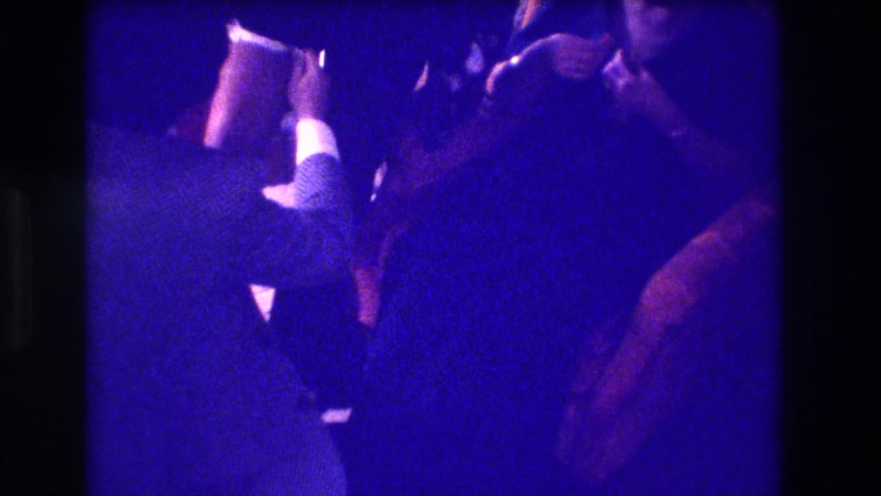 Blue-saturated 8mm frame from the 1973 Medford clip with adults leaning close in a private interior.