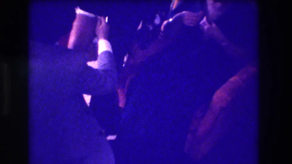 Blue-saturated 8mm frame from the 1973 Medford clip with adults leaning close in a private interior.