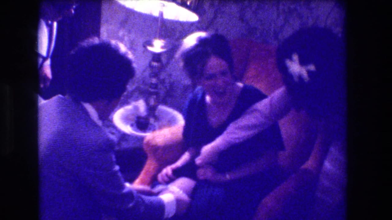 Two adults seated close together indoors; a man in a light jacket leans toward a laughing woman in a dark top, under a round lamp.