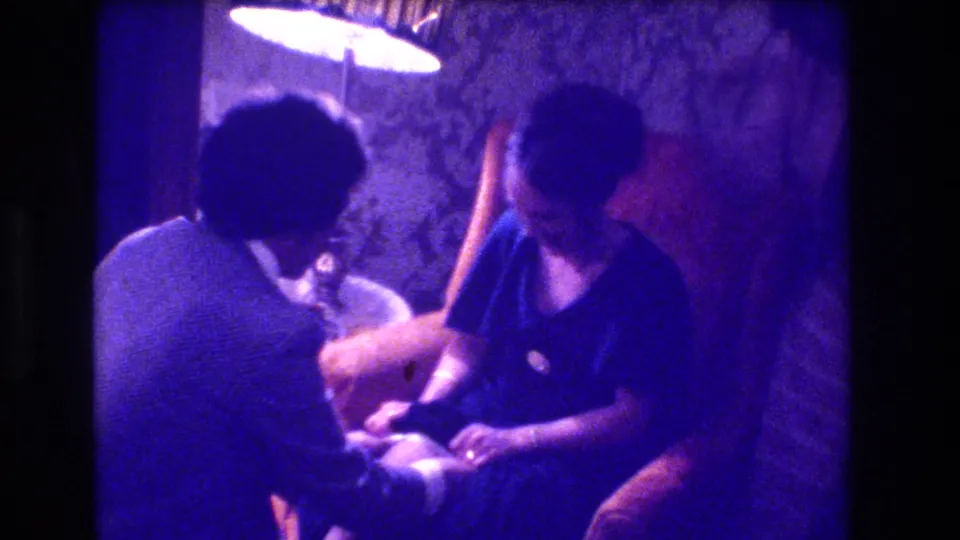 8mm home movie, circa 1973, showing a man's back as he sits with a woman in a dark top in a warmly lit domestic interior with purple-blue color shift.
