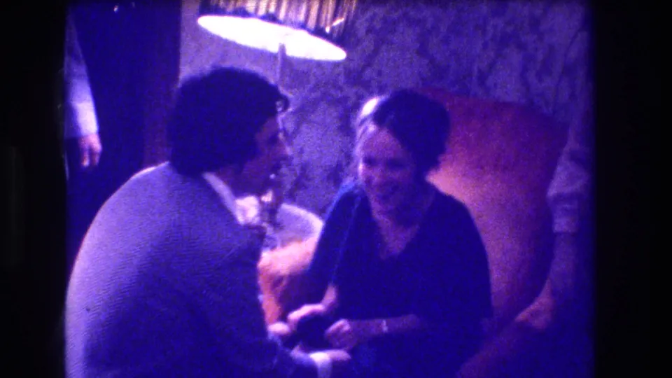 8mm home movie, circa 1973, Medford New Jersey, showing a smiling woman in a dark dress beside a man in a lighter jacket under a round lamp.