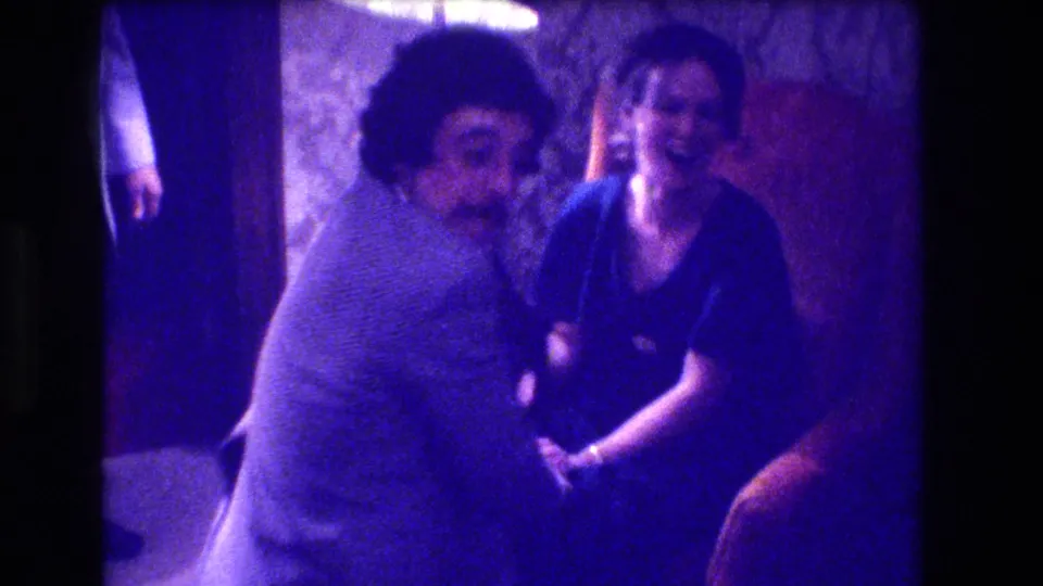 8mm home movie, circa 1973, showing a man and woman smiling toward the camera in a domestic interior with textured wallpaper and a wooden decorative element behind them.